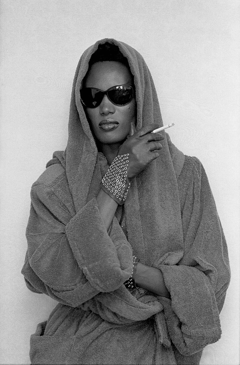 Grace Jones wearing sunglasses and a hooded coat, holding a cigarette in Los Angeles in 1984.