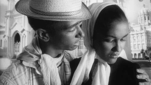 A black-and-white film still with two people standing side by side, one wearing a hat.