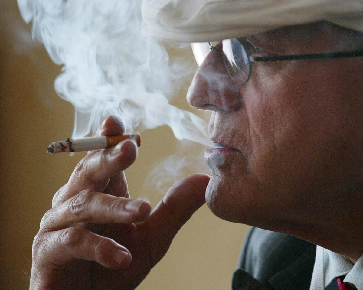 A man in glasses holding a cigarette, smoke drifting across his face.