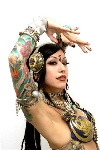 A tattooed woman wearing gold headphones, layered jewelry, and a patterned top.