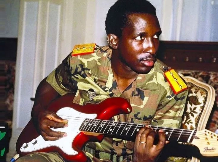 Thomas Sankara seated with a red electric guitar, wearing a camouflage uniform.